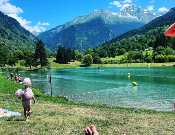 Vue en POV du lac de Bozel depuis la rive de la baignade surveillée, avec des reflets scintillants sur l’eau et un paysage de montagne en arrière-plan.