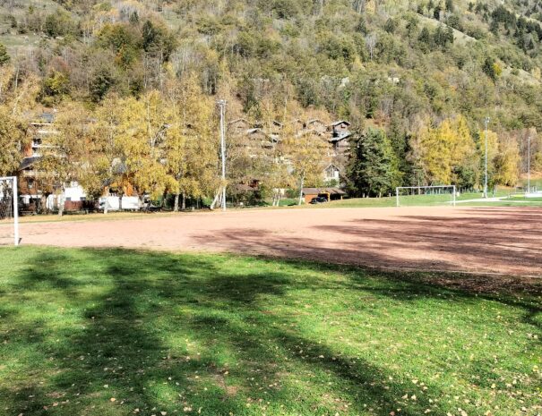 Terrain de football en terre battue à Bozel, entouré de verdure et de montagnes, sous un ciel dégagé.